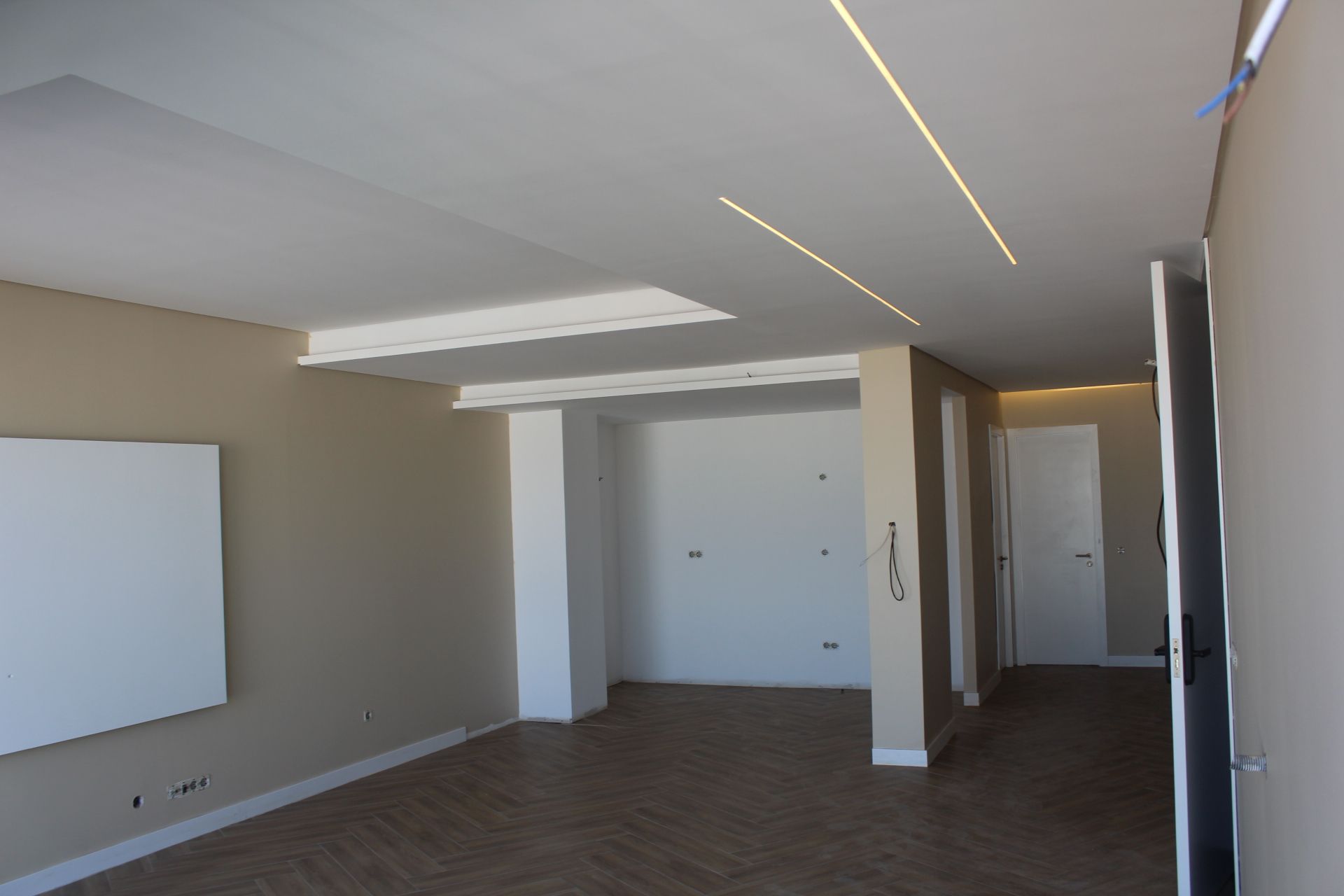 Sala vazia com luzes embutidas no teto, paredes bege e piso com aparência de madeira.