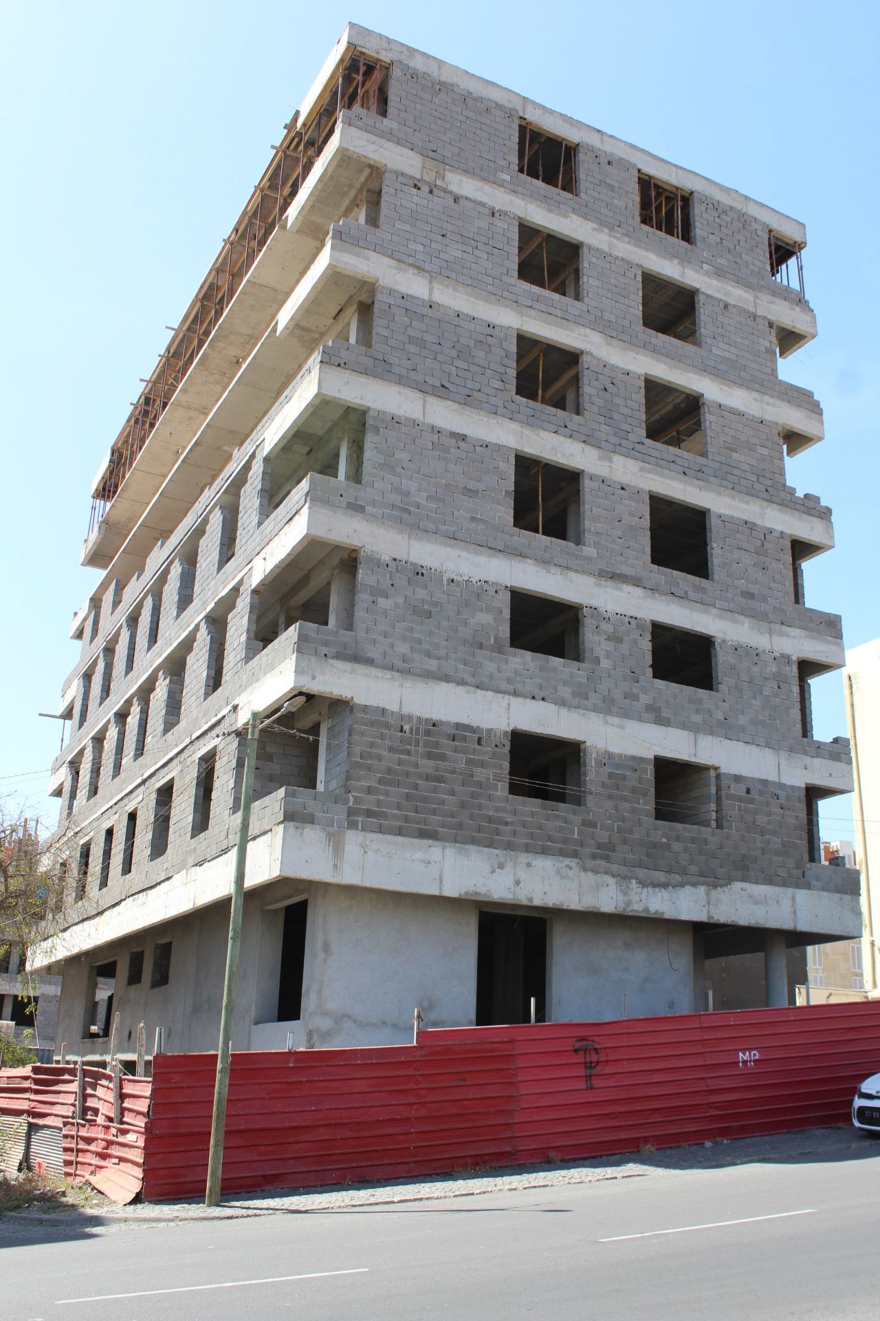 Prédio em construção, estrutura de concreto cinza, seis andares de altura, cerca vermelha na frente.