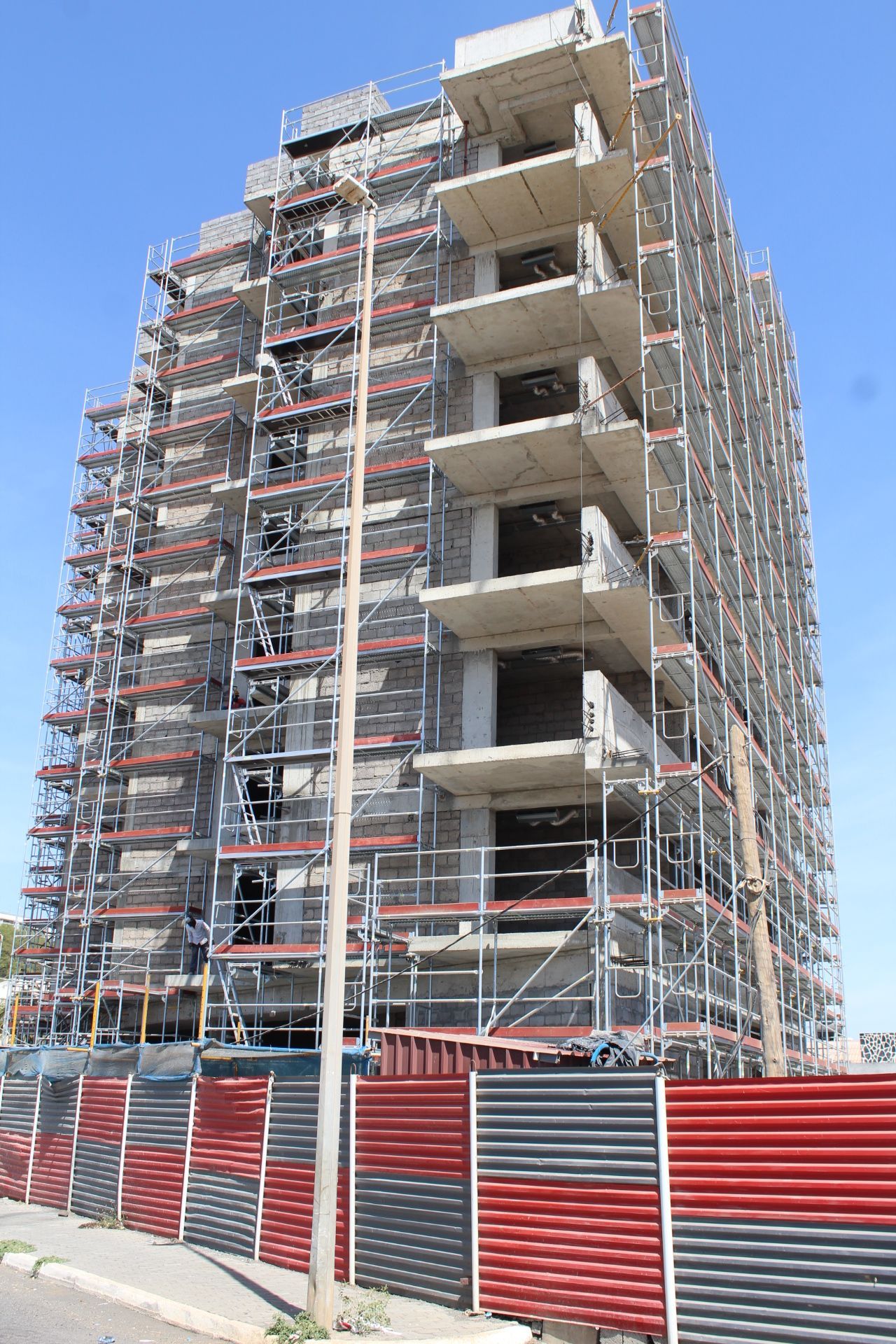 Prédio em construção, cercado por andaimes e cercas vermelhas/cinzas, contra um céu azul.
