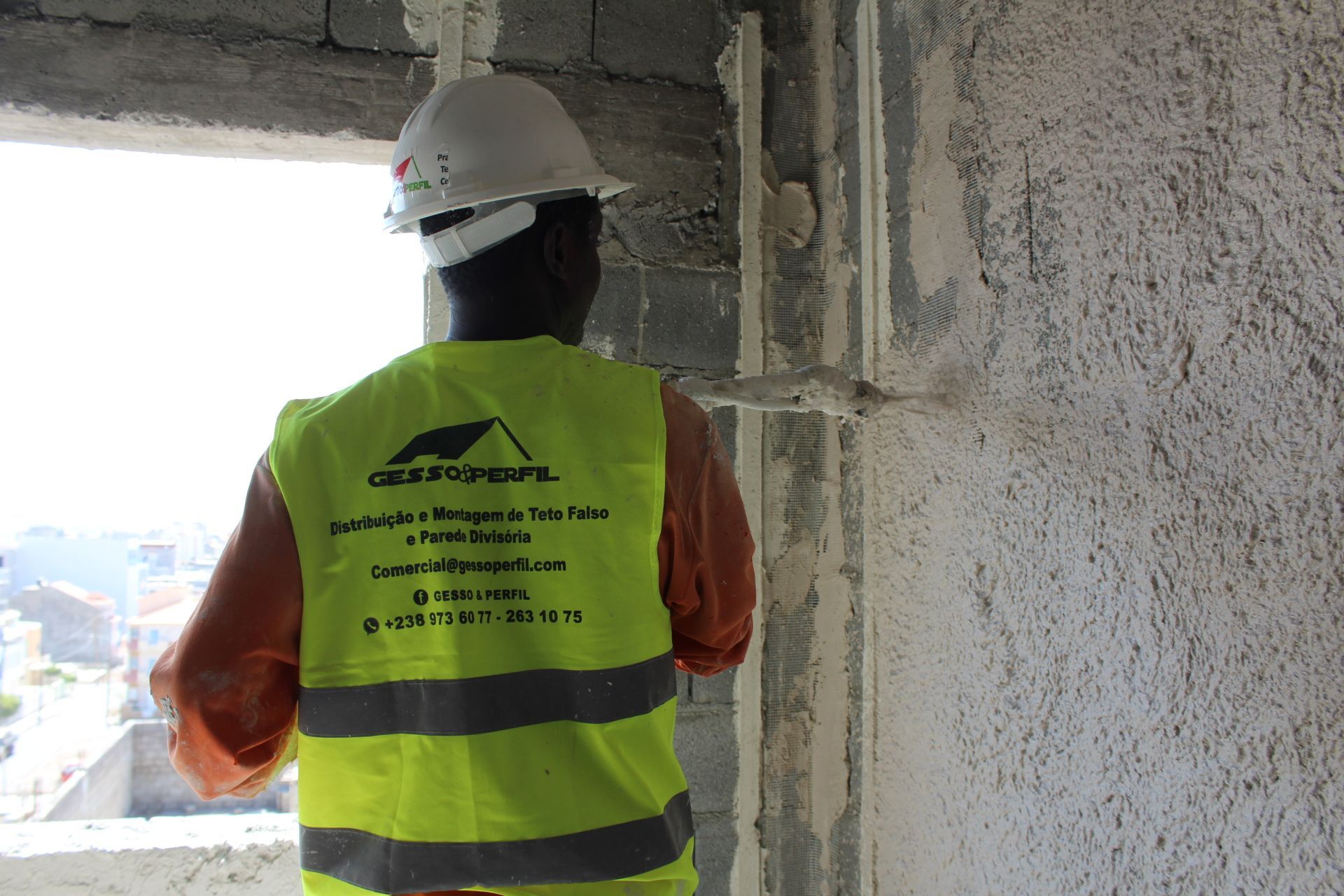Trabalhador da construção civil usando capacete e colete de segurança, inspecionando um canto de prédio com uma janela.