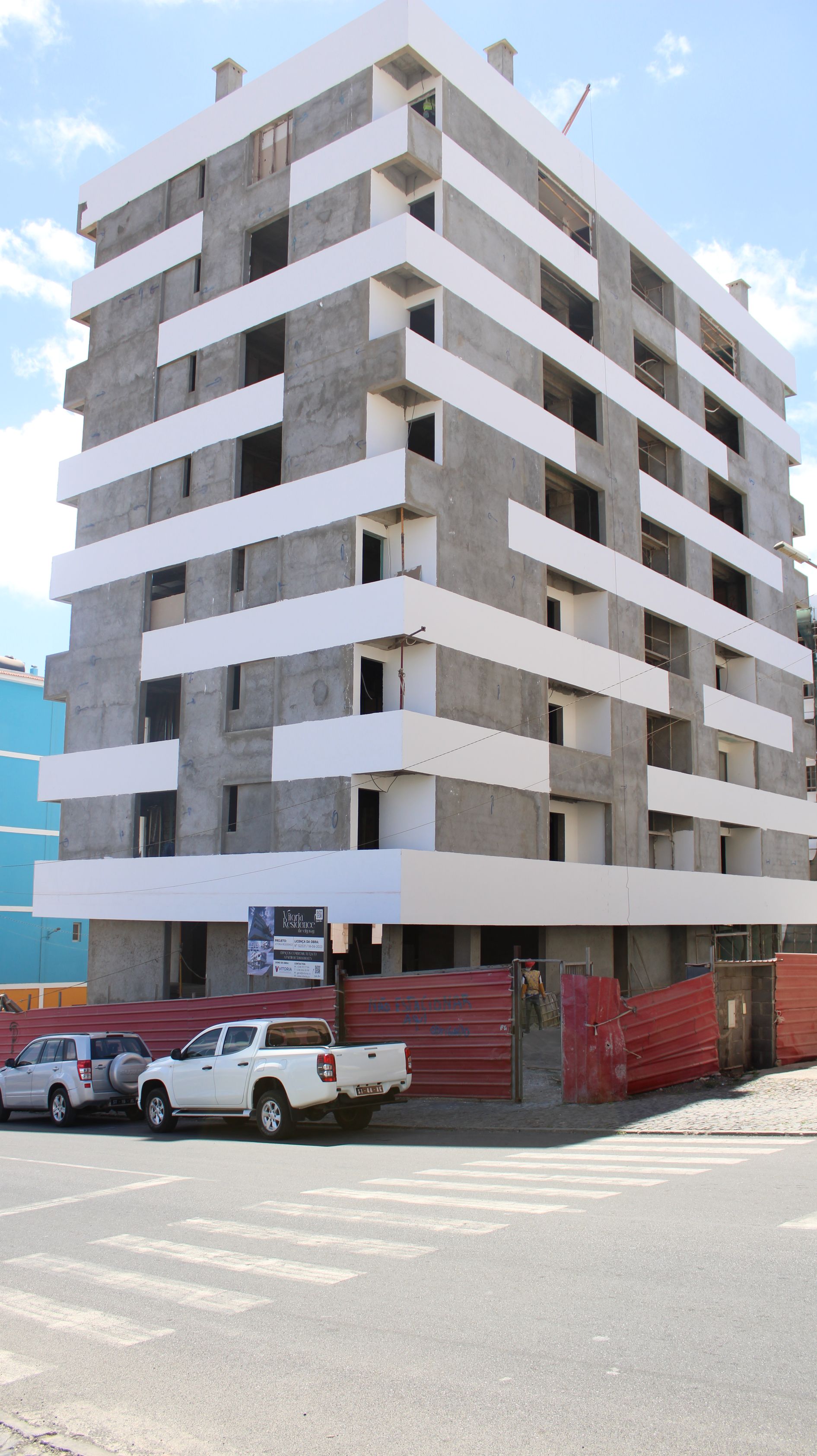 Prédio de vários andares em construção; faixas horizontais brancas, concreto cinza, barreira de construção vermelha.