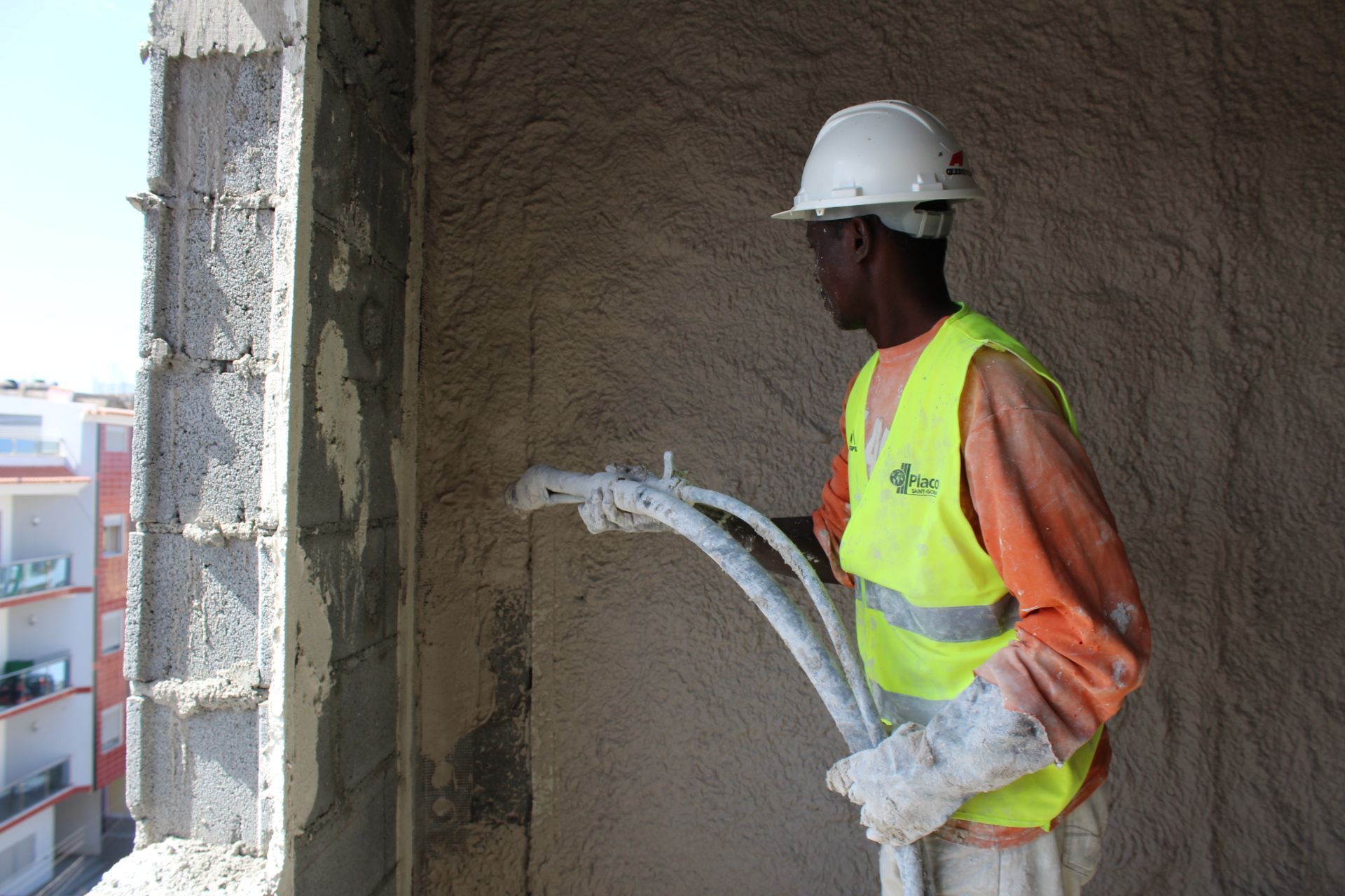 Trabalhador da construção civil pulverizando gesso na parte externa de um prédio, usando colete de segurança e capacete.