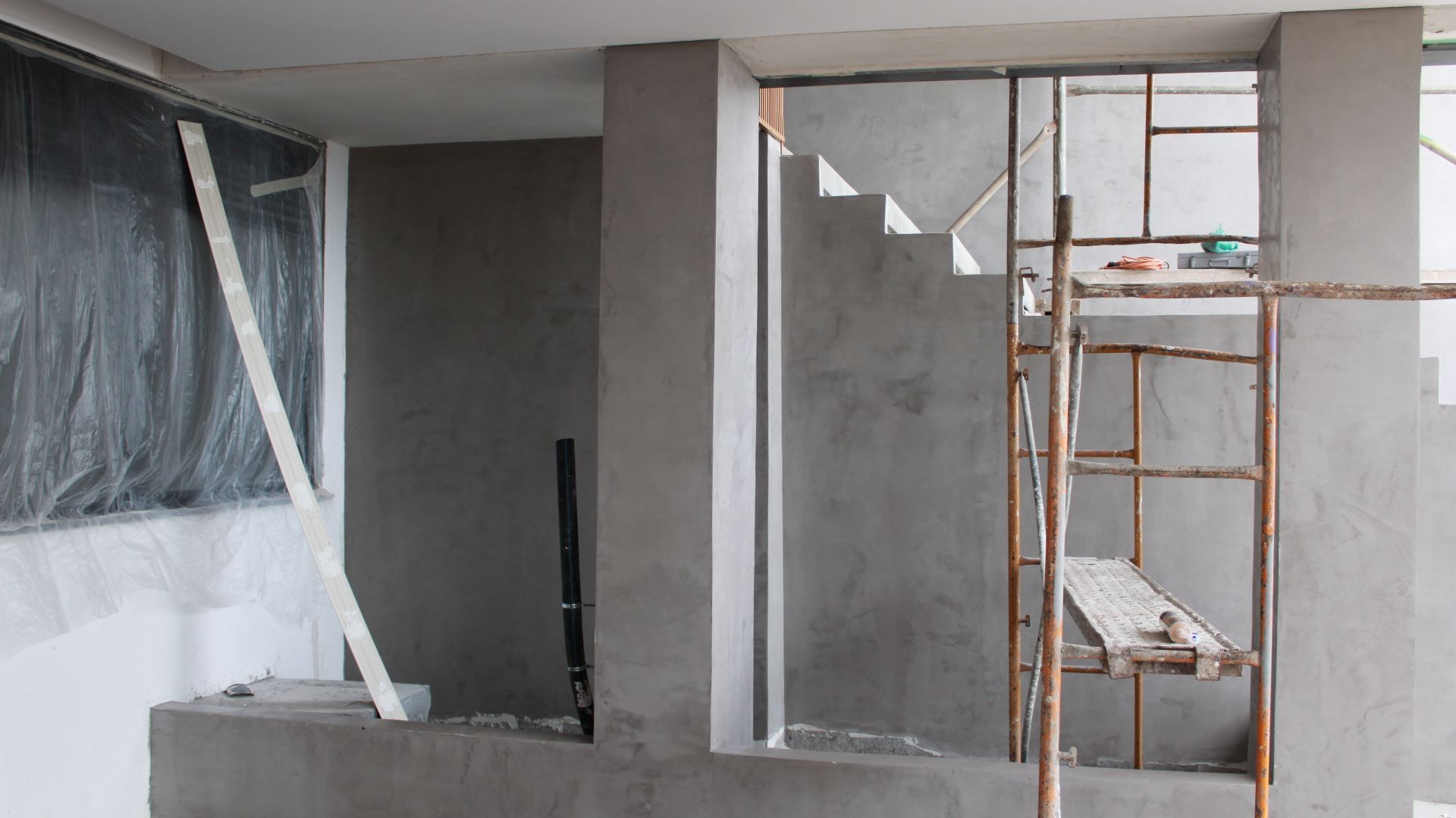 Canteiro de obras com escadas inacabadas, paredes cinzas e um andaime de madeira.