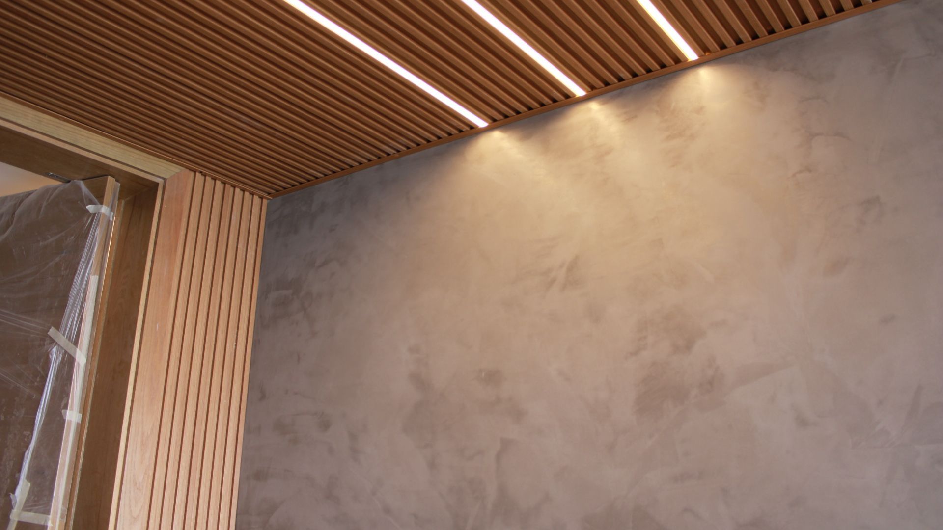 Teto de ripas de madeira com iluminação embutida iluminando uma parede texturizada em tom bege.