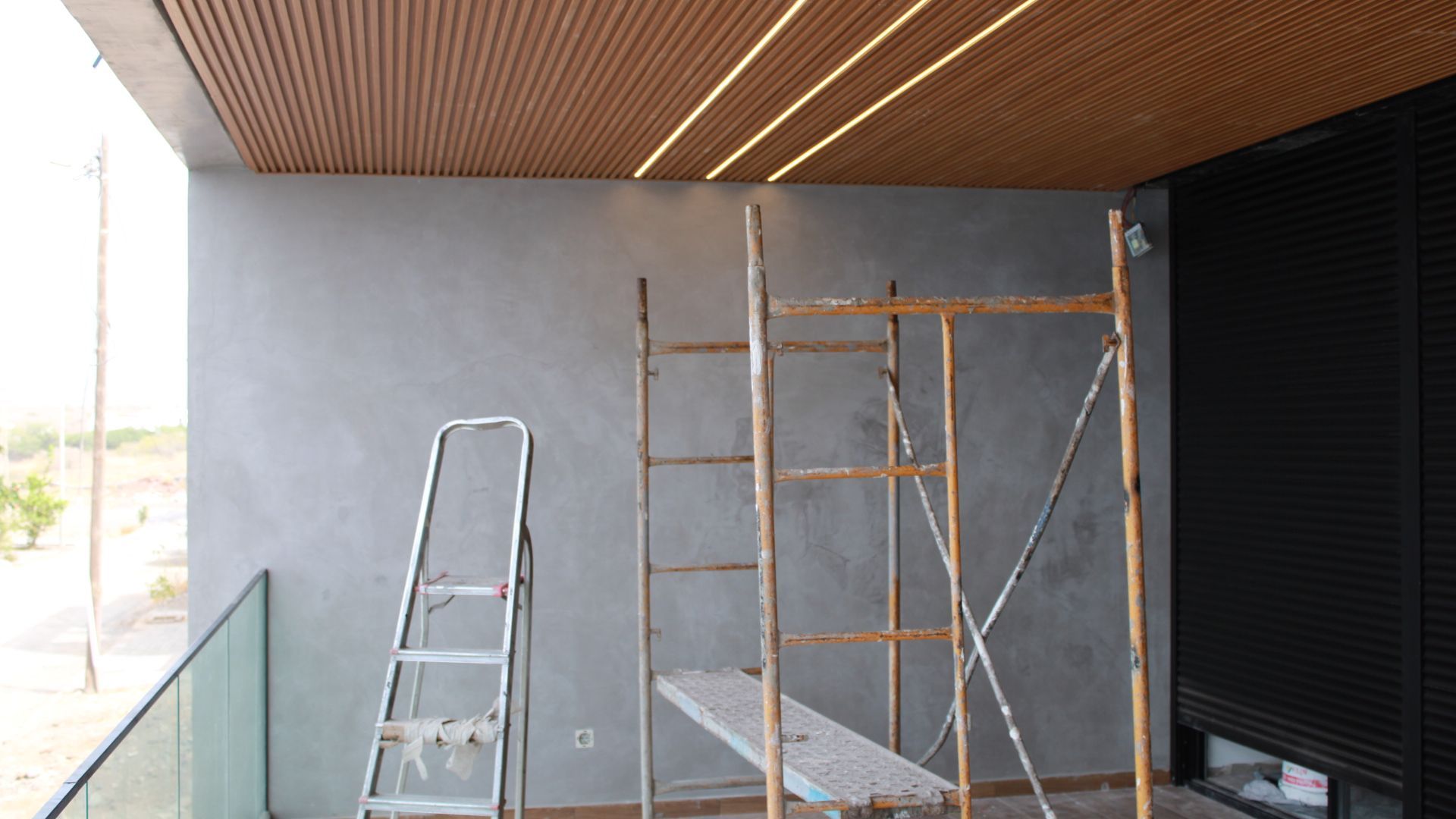 Uma varanda com parede texturizada cinza, teto de madeira com luzes embutidas e andaimes.