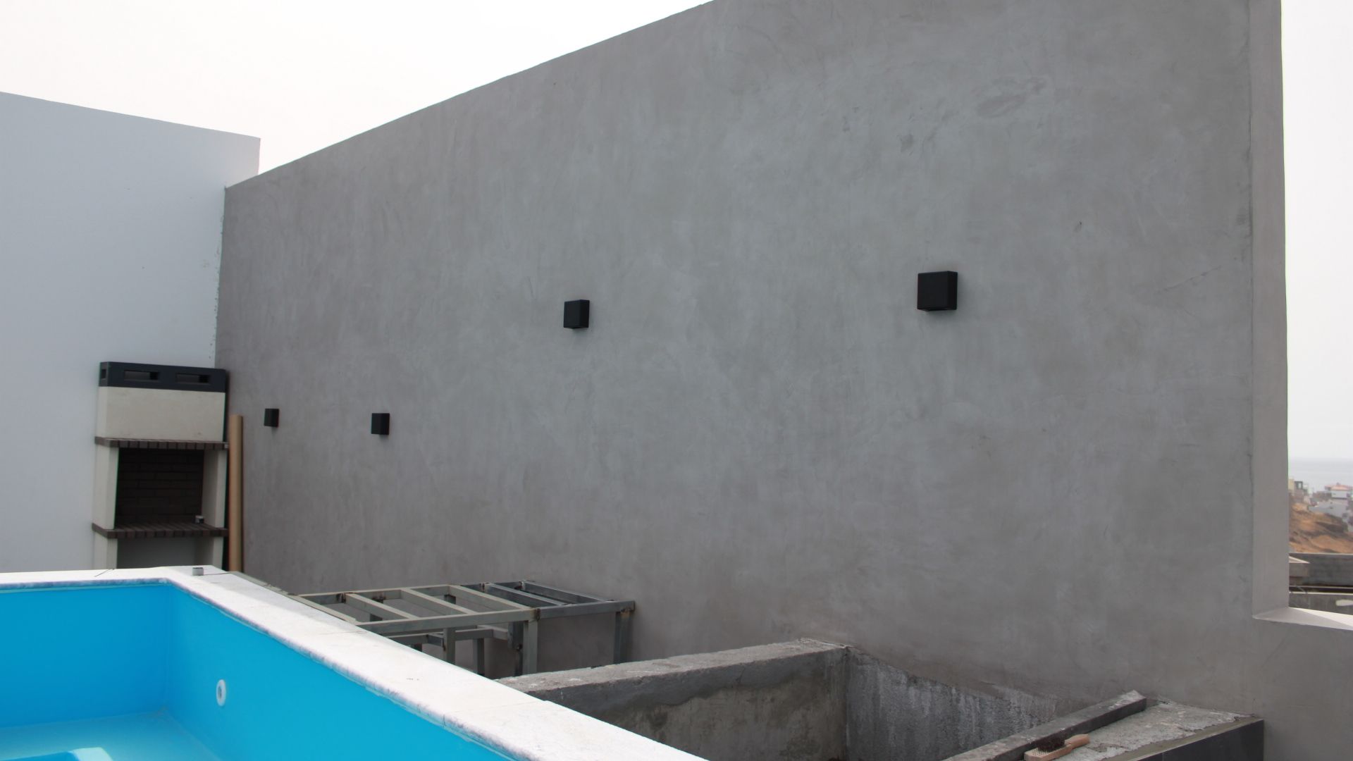 Parede cinza com luzes pretas quadradas, ao lado de uma piscina e área de churrasco, em um terraço.