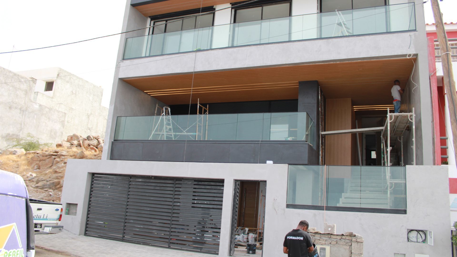 Prédio cinza moderno em construção, varandas de vidro, trabalhadores presentes.