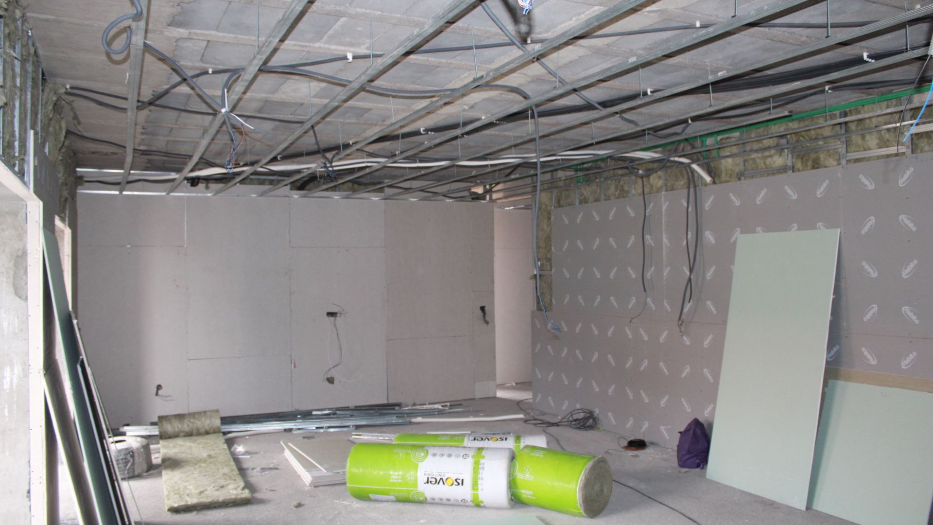 Interior do canteiro de obras: paredes e estrutura do teto visíveis, rolos de isolamento e placas de drywall presentes.