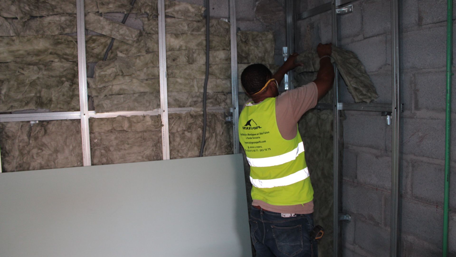 Pessoa instalando isolamento entre montantes metálicos; canteiro de obras. Usando colete e óculos de segurança.