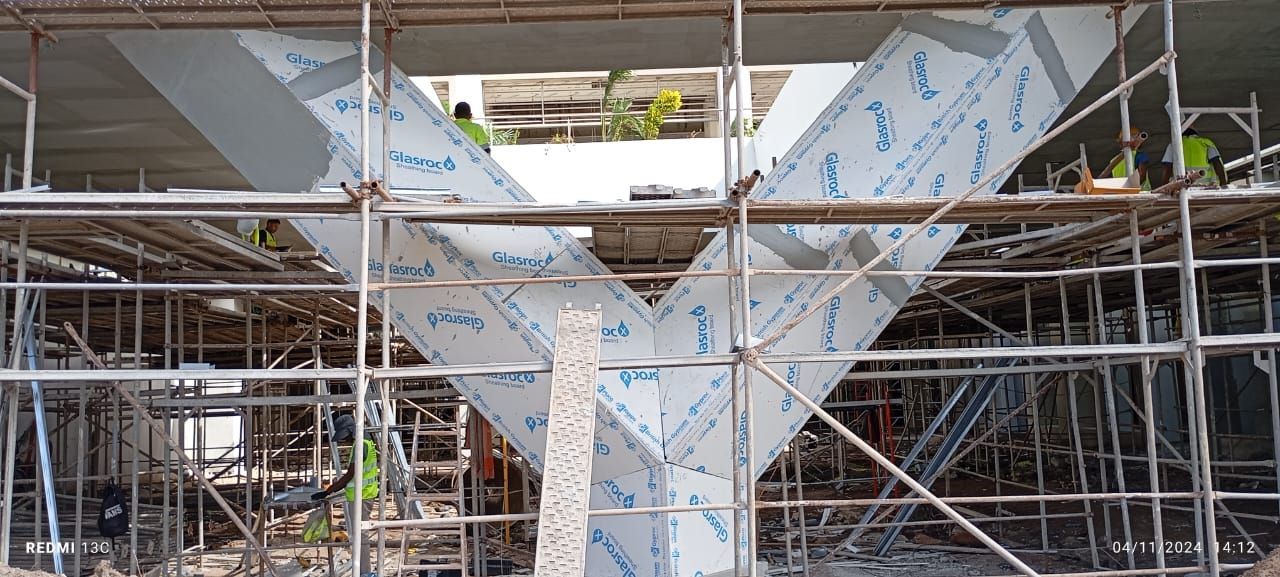 Canteiro de obras com andaimes e estruturas metálicas, algumas com película protetora, e com presença de trabalhadores.