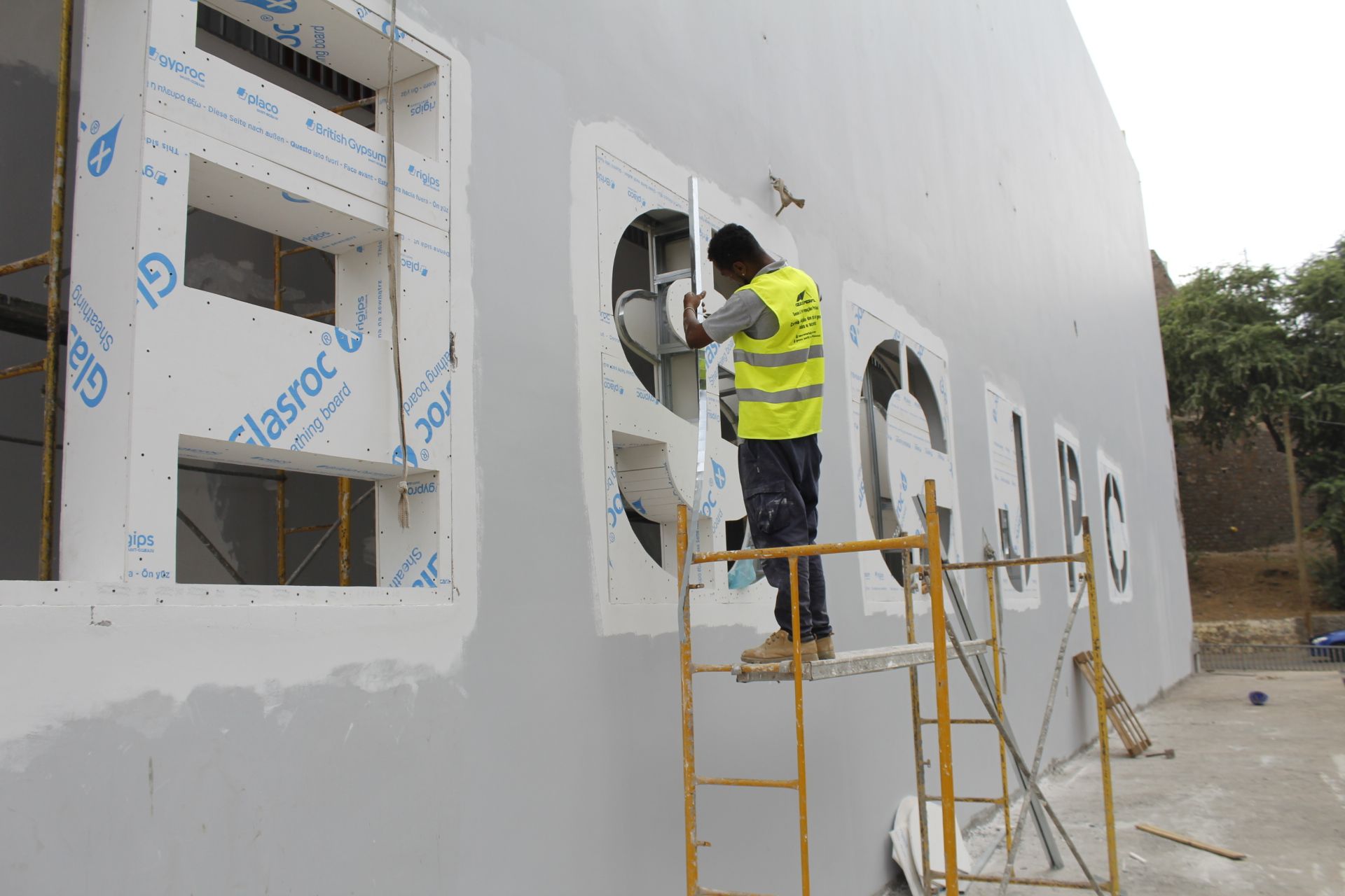 Trabalhador da construção civil com colete de segurança em andaime instalando painéis na fachada de um edifício.