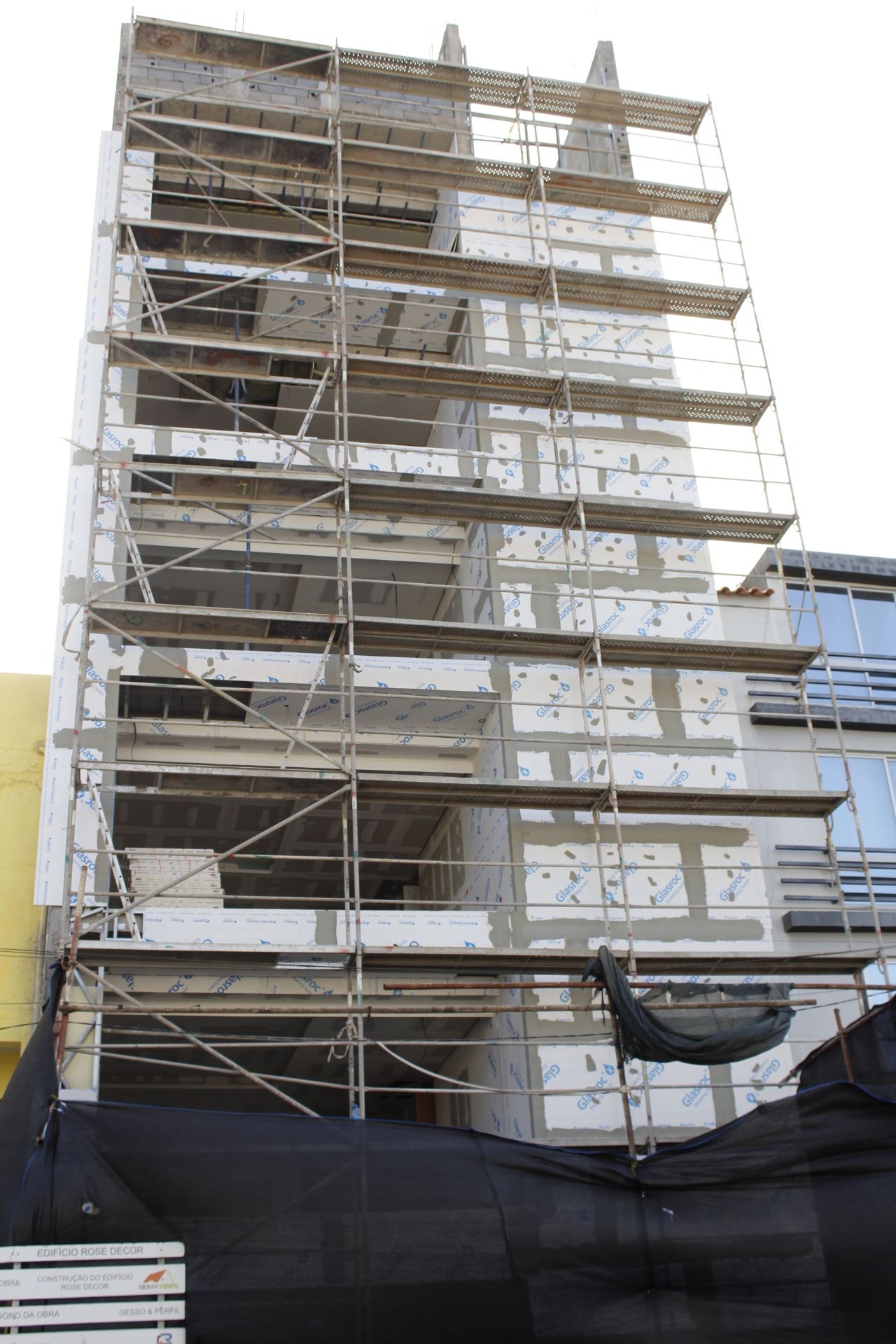 Prédio em construção com andaimes. Fachada cinza e branca, andares visíveis.