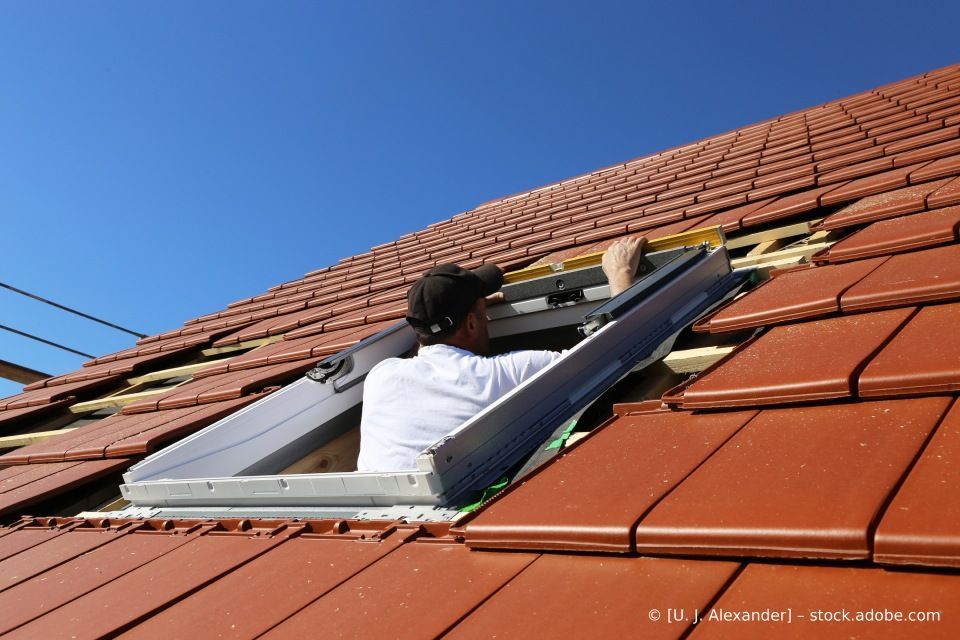 Arbeiter repariert auf einem roten Ziegeldach ein Dachfenster unter klarem, blauem Himmel