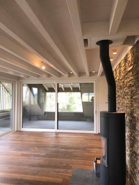 Ein Zimmer mit Holzböden, freiliegenden weissen Deckenbalken, einer Steinwand & einem schwarzen Holzofen an großen Fenstern.