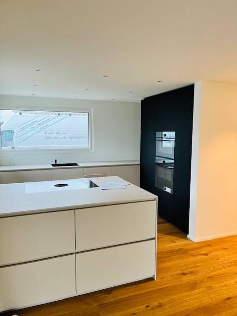 Moderne Küche mit hellgrauer Kochinsel, weissen Arbeitsplatten und schwarzem Wandschrank auf hellem Holzboden.