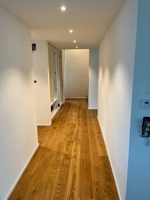 Ein schmaler Flur mit weissen Wänden und hellem Holzboden, ausgestattet mit Deckeneinbauleuchten und einem eingebauten Regal.