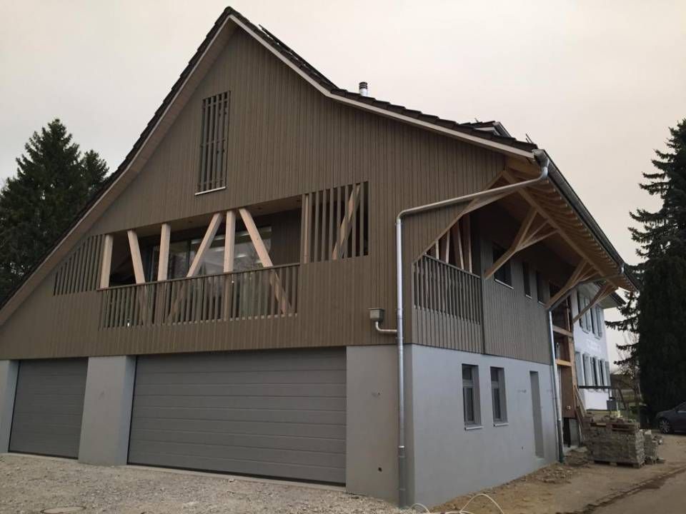 Ein braunes, holzverkleidetes Haus im Scheunenstil mit grauen Garagentoren und Betonfundament vor einem Hintergrund.