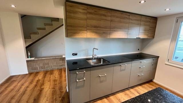 Eine Küche mit grauen Unterschränken, holzfarbenen Oberschränken, schwarzen Arbeitsplatten und einer Treppe im Hintergrund.