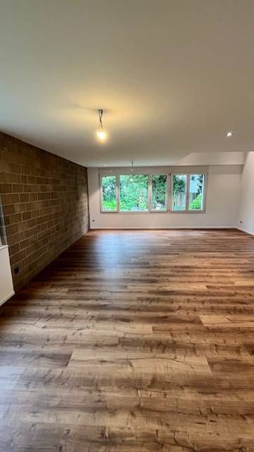Ein geräumiges, leeres Zimmer mit Holzdielenboden, einer braunen Backsteinwand auf der linken Seite und einer Fensterreihe.