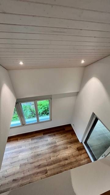 Ein Blick von oben in einen Raum mit Holzboden, weissen Wänden, einer schrägen Holzdecke und zwei grossen Fenstern.