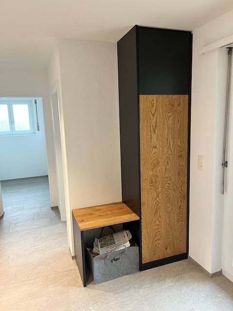 Ein schwarzes Aufbewahrungsmöbel mit Holzmaserung, bestehend aus einem hohen Schrankfach neben einer niedrigeren Sitzbank.