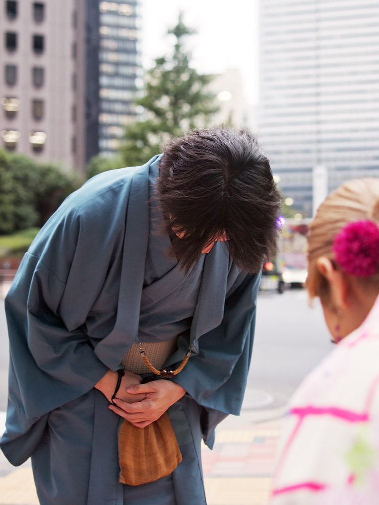 Une personne en kimono bleu s'inclinant devant une personne en kimono rose ; arrière-plan de la ville.