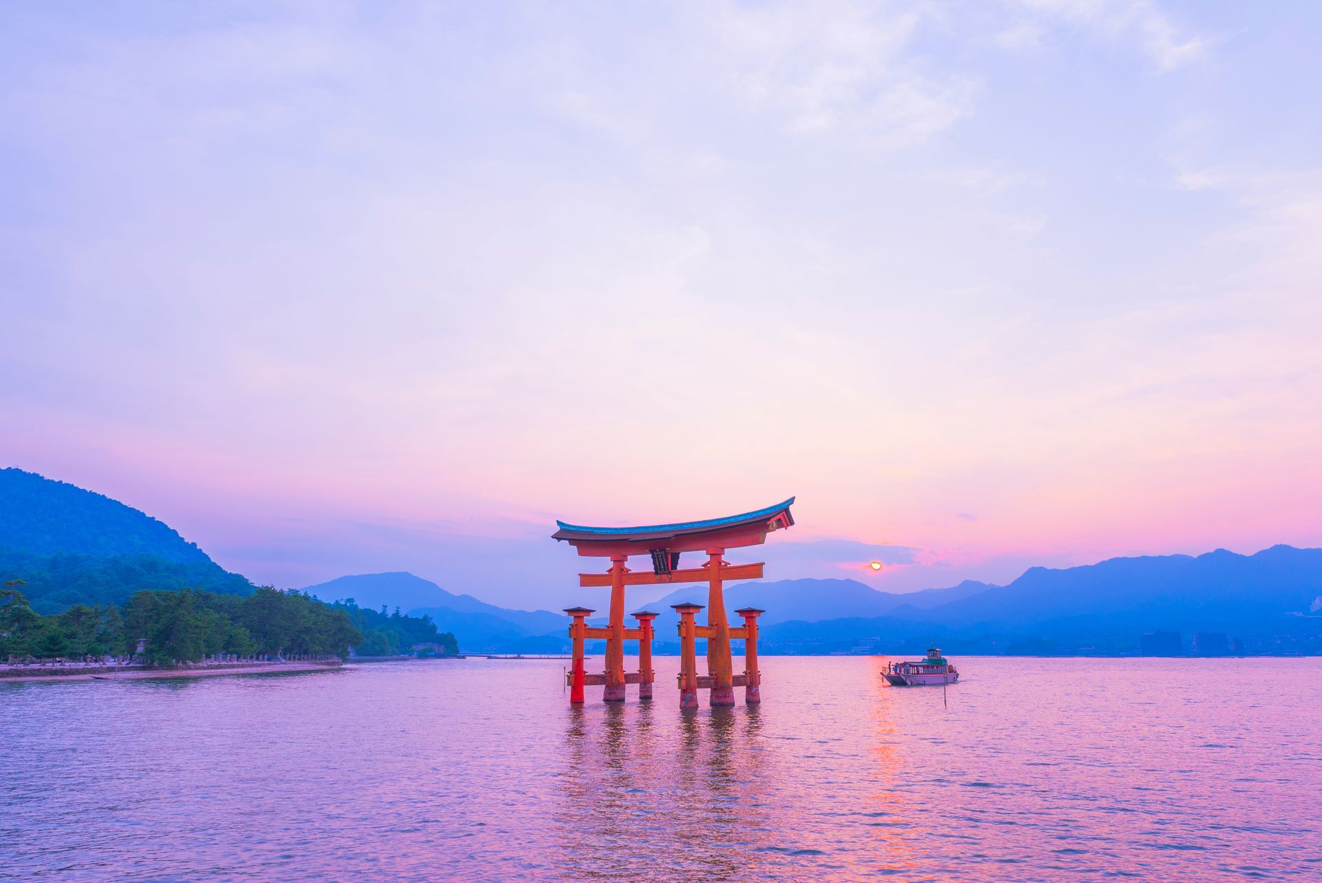Porte torii orange flottante dans l'eau au coucher du soleil, avec des montagnes au loin ; Miyajima, Japon.