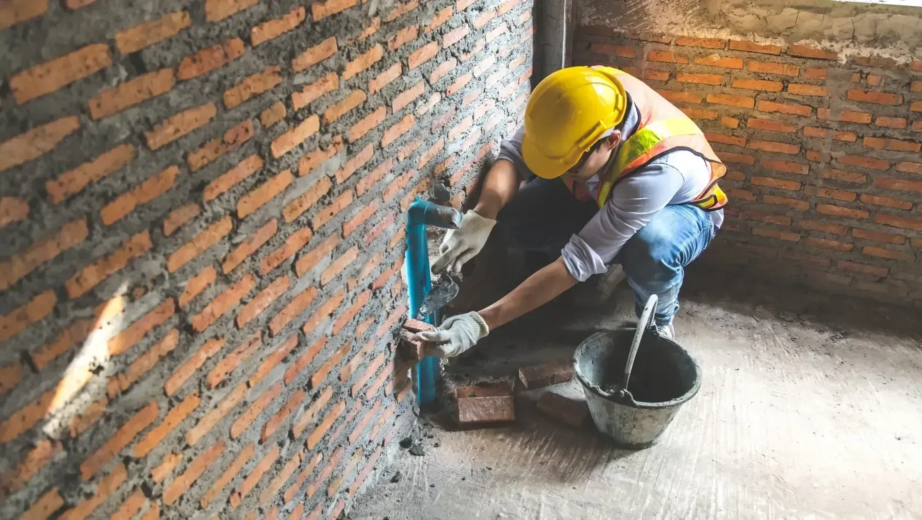 Obrero de la construcción instalando una tubería azul contra una pared de ladrillo, con casco y guantes.