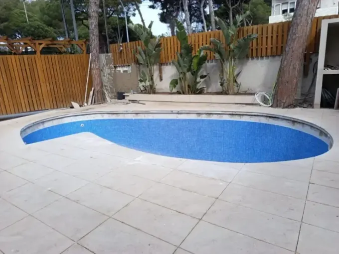 Piscina con azulejos azules rodeada de azulejos beige y una valla de madera; árboles y plantas al fondo.