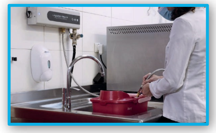 Persona con bata de laboratorio cerca de un fregadero. Sostiene una herramienta y trabaja con un recipiente rojo.