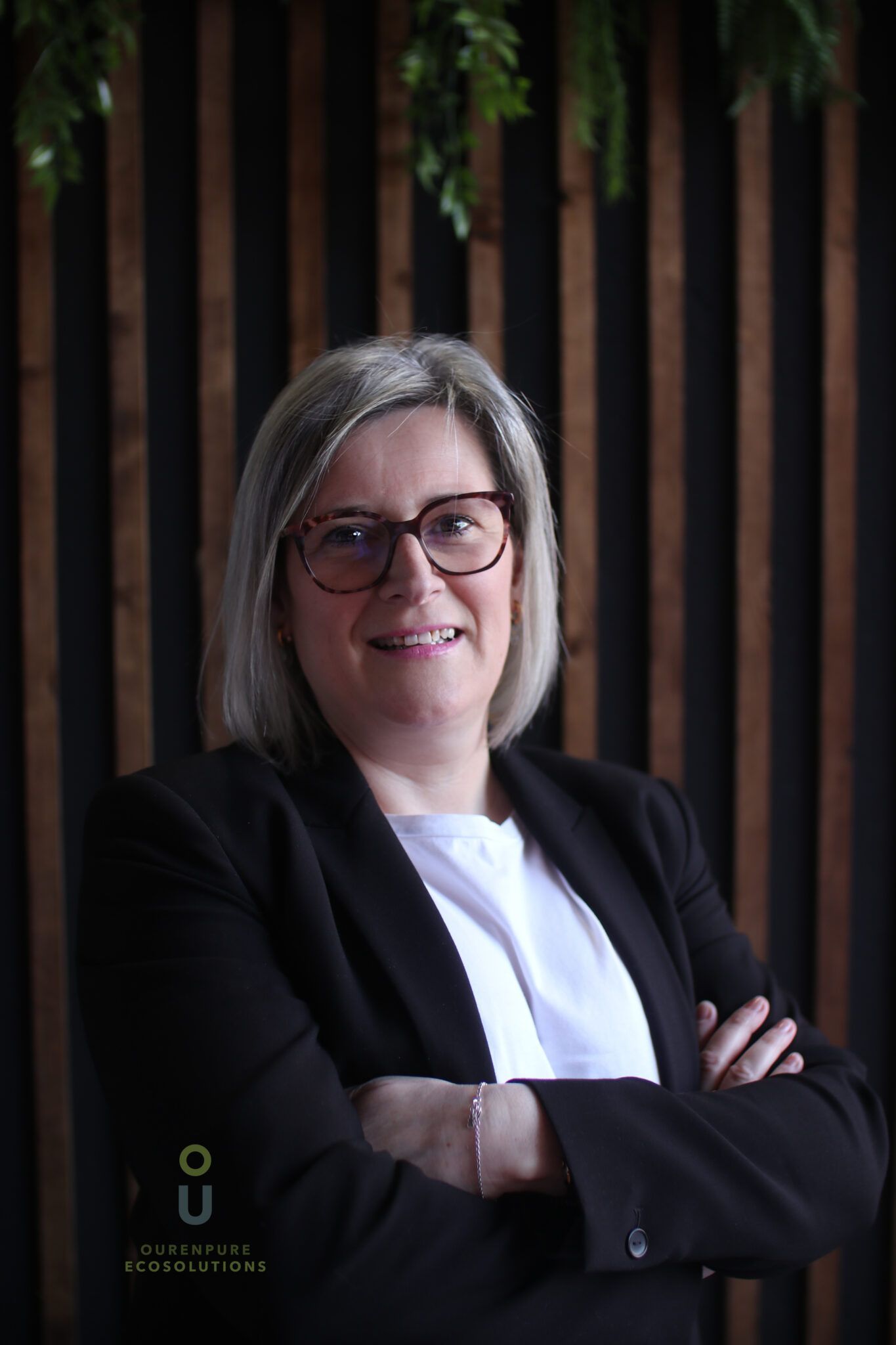 Mujer con gafas y blazer negro, con los brazos cruzados, sonriendo. Fondo de madera oscura.