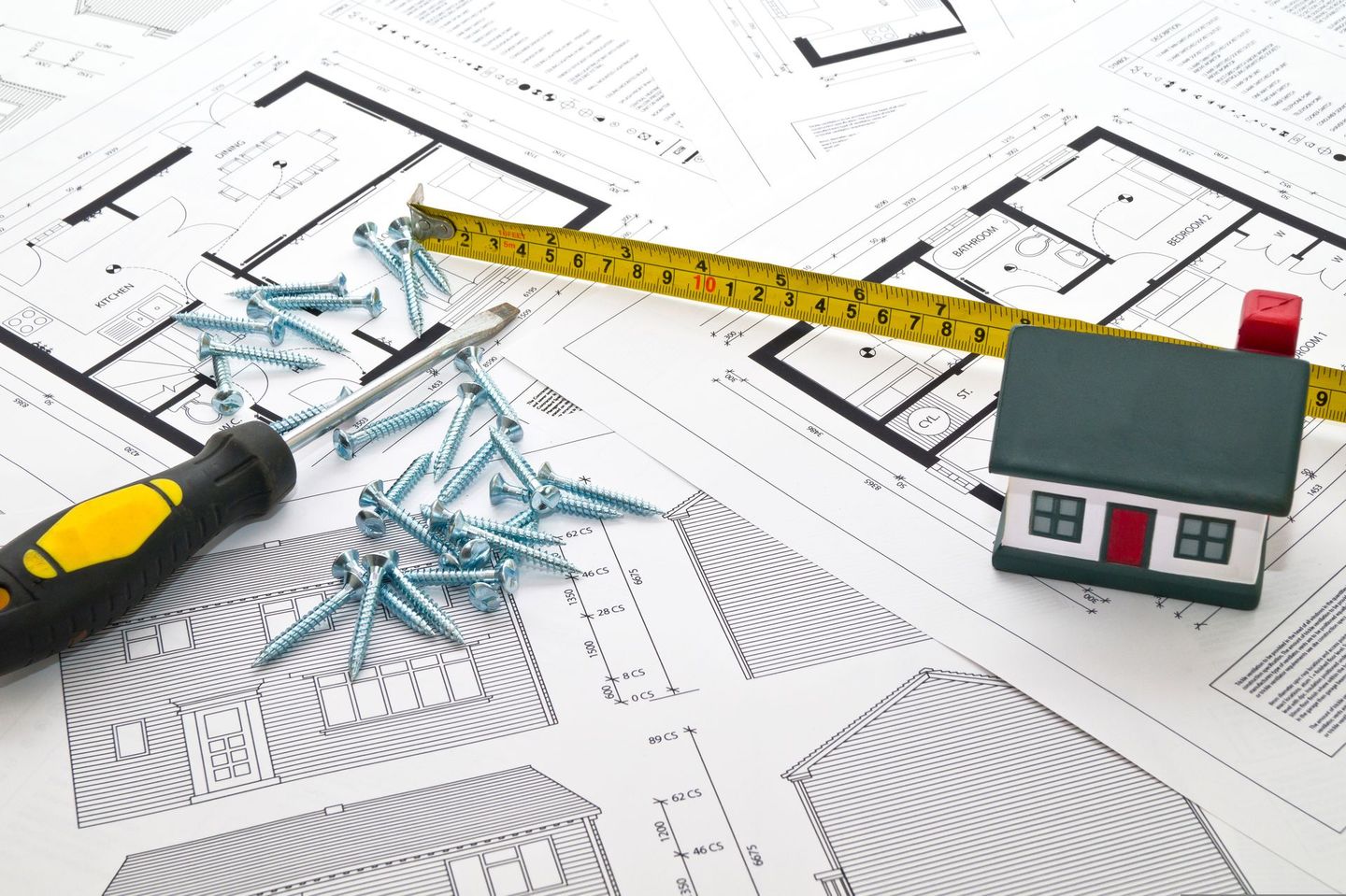 Una casa modelo está colocada encima de un plano junto a una cinta métrica y tornillos.