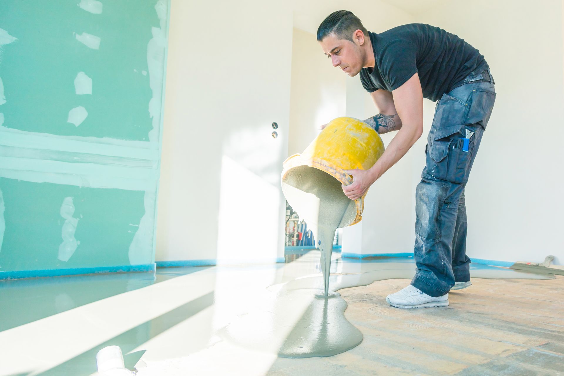 Mann gießt Beton aus einem gelben Eimer auf den Boden eines Raums mit teilweise fertigen Wänden.