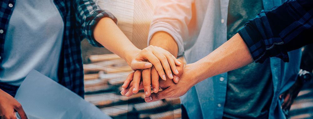 Die Hände sind in einer Team-Gruppe übereinander gestapelt, im Sonnenlicht ist ein Bauplan zu sehen.