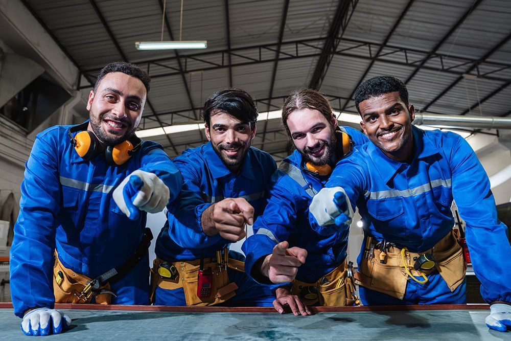 Vier Arbeiter in blauen Overalls zeigen in einer Werkstatt lächelnd auf den Betrachter.