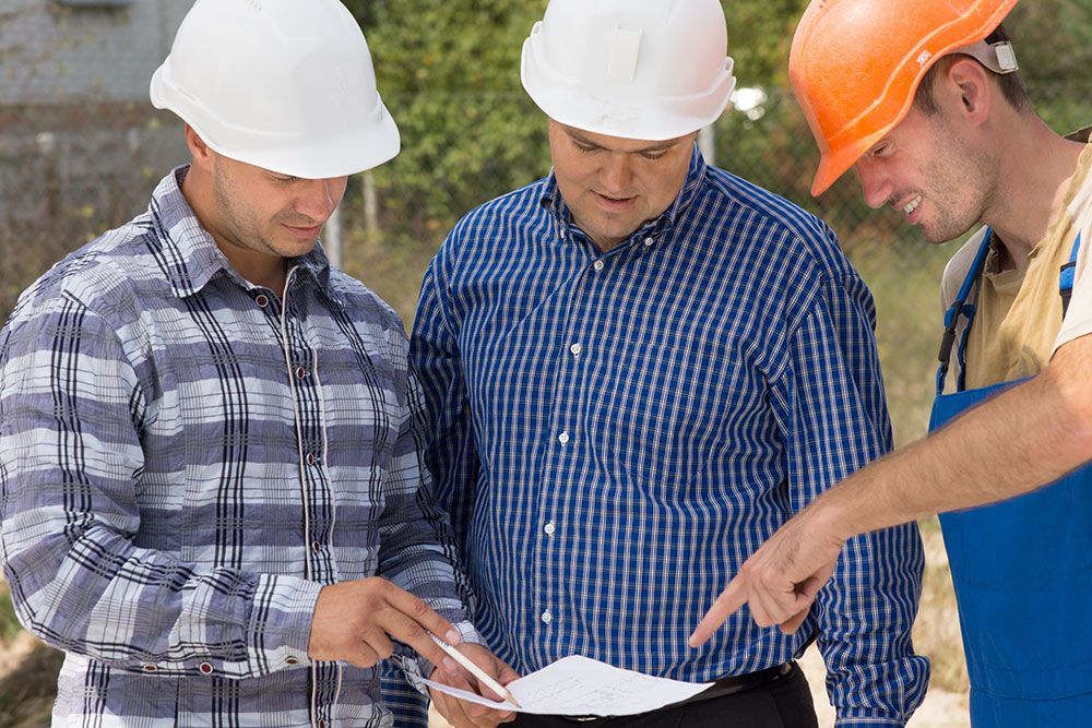 Drei Bauarbeiter überprüfen auf einer Baustelle Baupläne, tragen Schutzhelme und zeigen mit dem Finger.