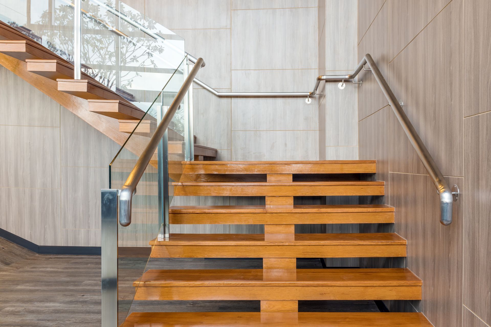 Holztreppe mit Glasgeländer und Metallhandläufen vor einer hellen holzgetäfelten Wand.