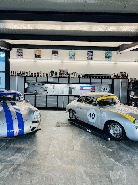 Deux voitures de course argentées d'époque dans un garage. L'une a des bandes bleues, l'autre une bande jaune.
