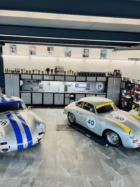 Deux Porsche de course classiques argentées dans un garage, l'une avec des bandes bleues, l'autre avec des bandes jaunes.