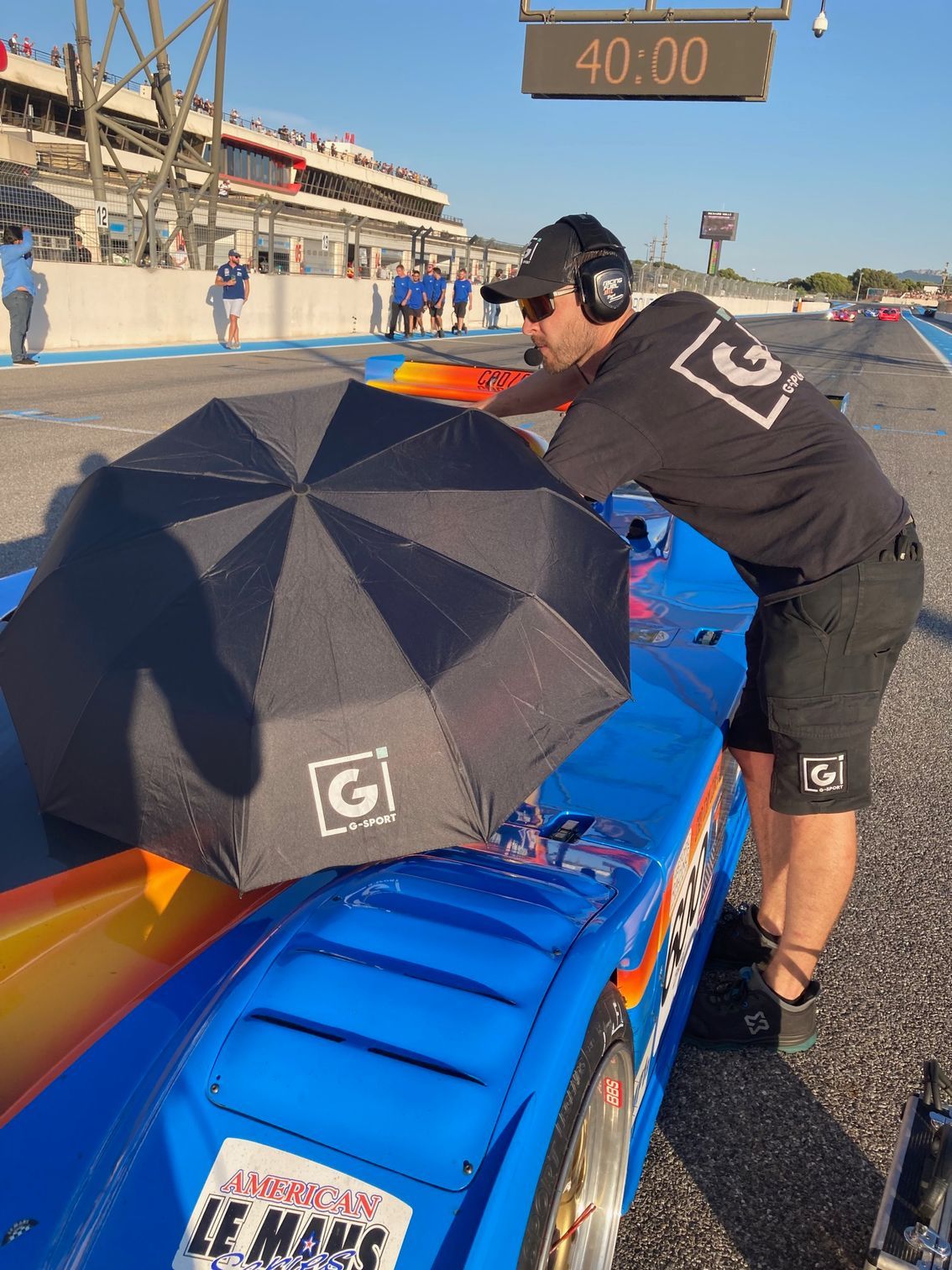 Un membre d'équipage, muni d'un parapluie, protège une voiture de course bleue dans la voie des stands.