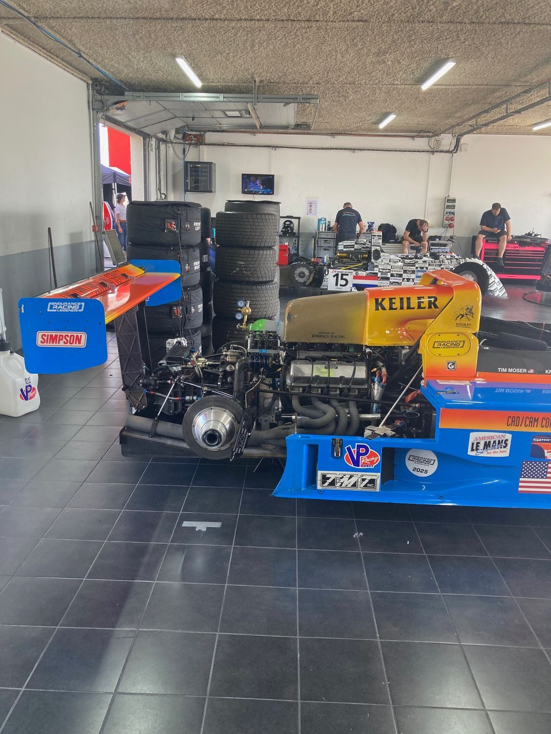 Voiture de course dans un garage, moteur visible, pneus empilés et mécaniciens.