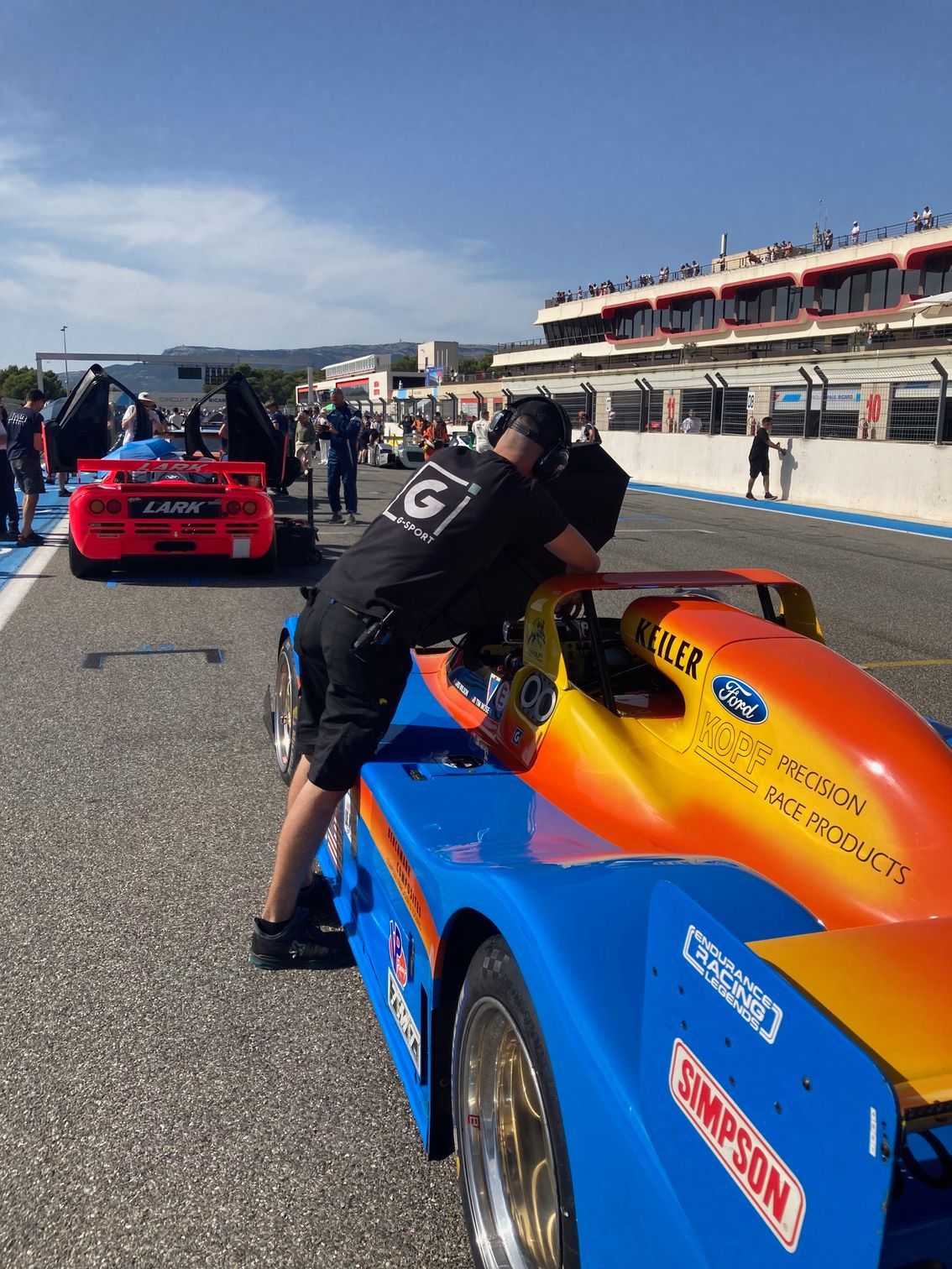 Un mécanicien travaille sur une voiture de course bleue et orange sur un circuit.