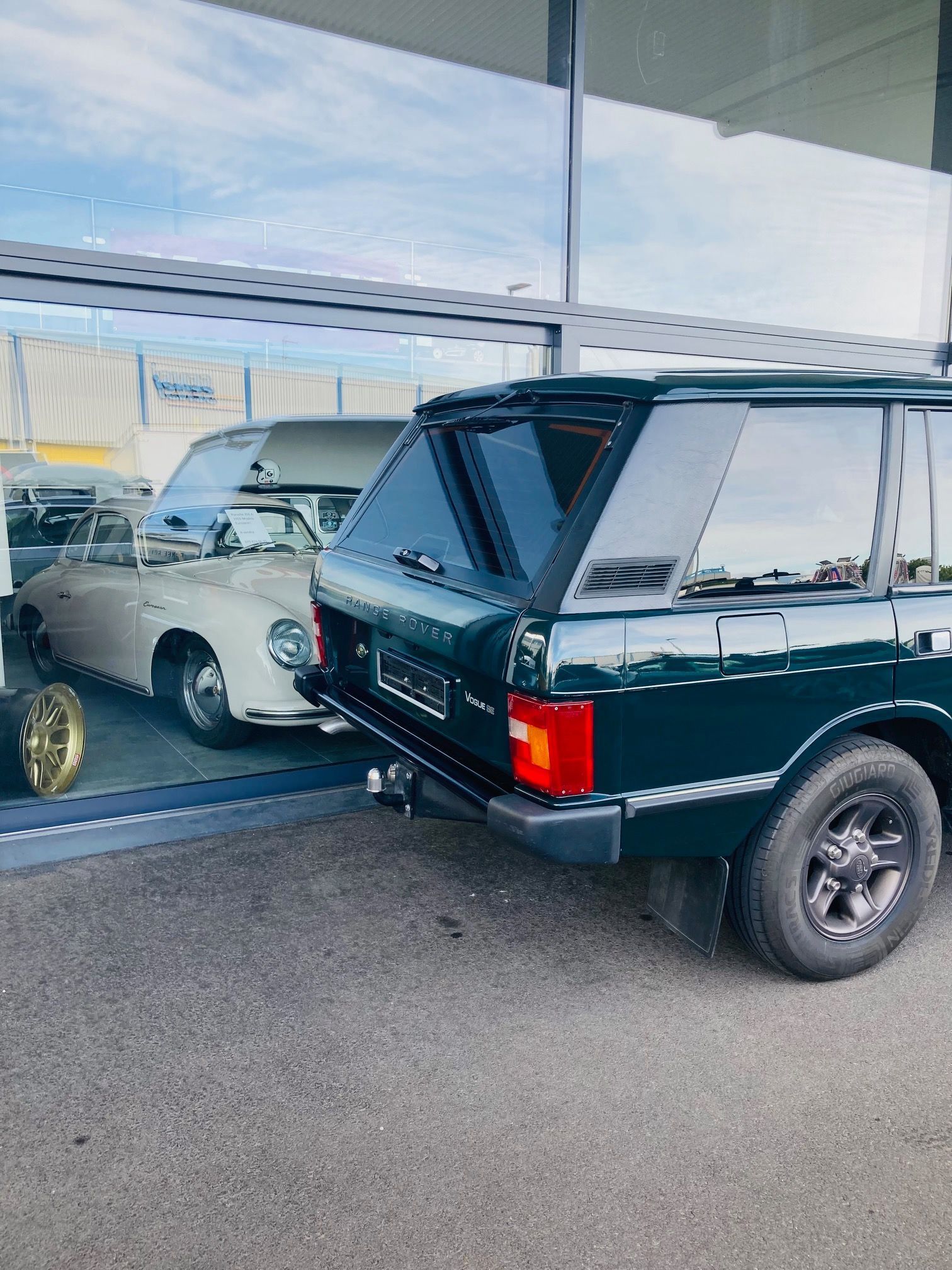 Un Range Rover vert est garé à côté d'une voiture classique de couleur claire, derrière une vitrine.