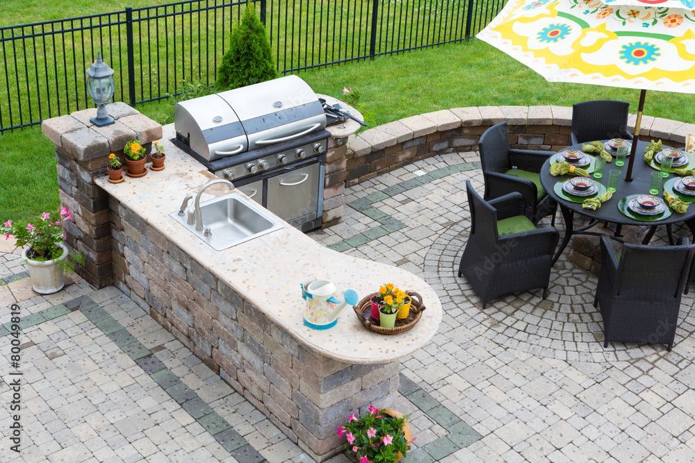 Patio sul retro con cucina esterna in mattoni, barbecue, tavolo da pranzo e sedie nere su pavimentazione.