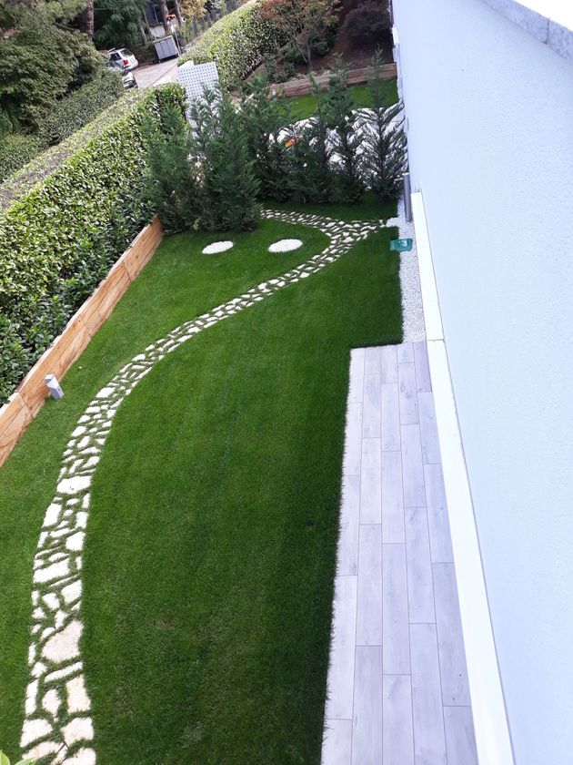 Giardino collinare paesaggistico con barriera nera contro le erbacce, rocce, erba verde e un tronco d'albero come elemento centrale.