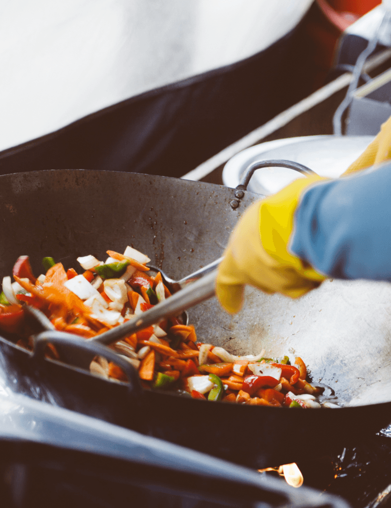 Eine Person mit gelben Handschuhen rührt Gemüse in einem Wok