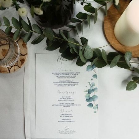 Auf einem Tisch liegt eine Speisekarte neben einer Blumenvase und einer Kerze.