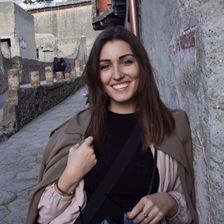 Una mujer de cabello castaño sonríe mientras posa en un antiguo callejón de piedra, vistiendo una camisa negra y una chaqueta