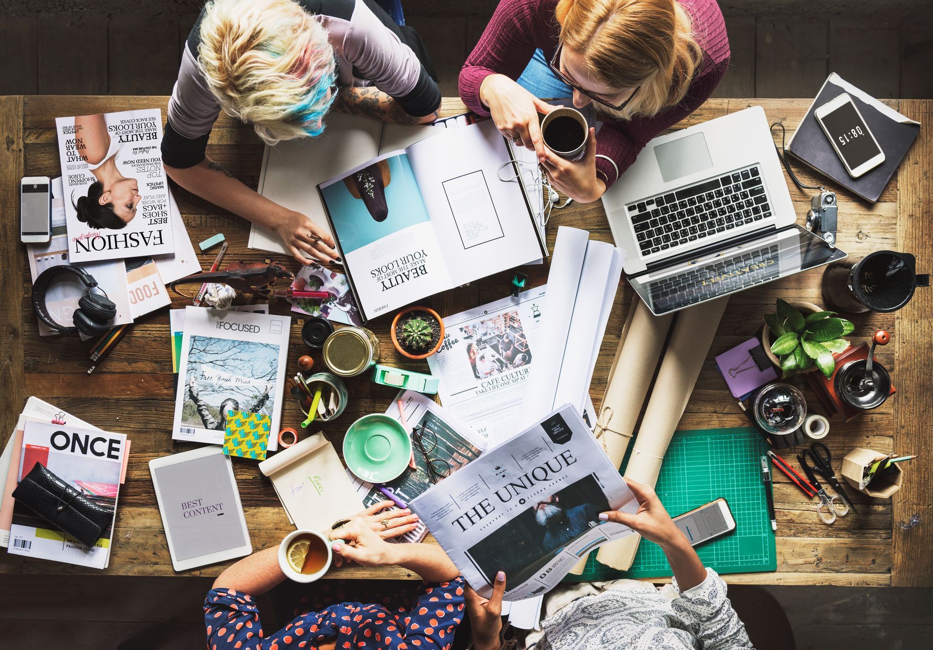 Tres personas en una mesa con portátiles, revistas y café, intercambiando ideas. Vista desde arriba.