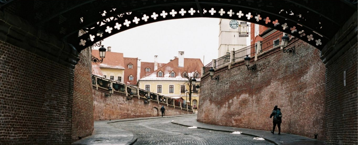 Vista de una calle histórica, enmarcada por un arco. Edificios con tejados de tejas rojas y una torre de reloj a lo lejos.
